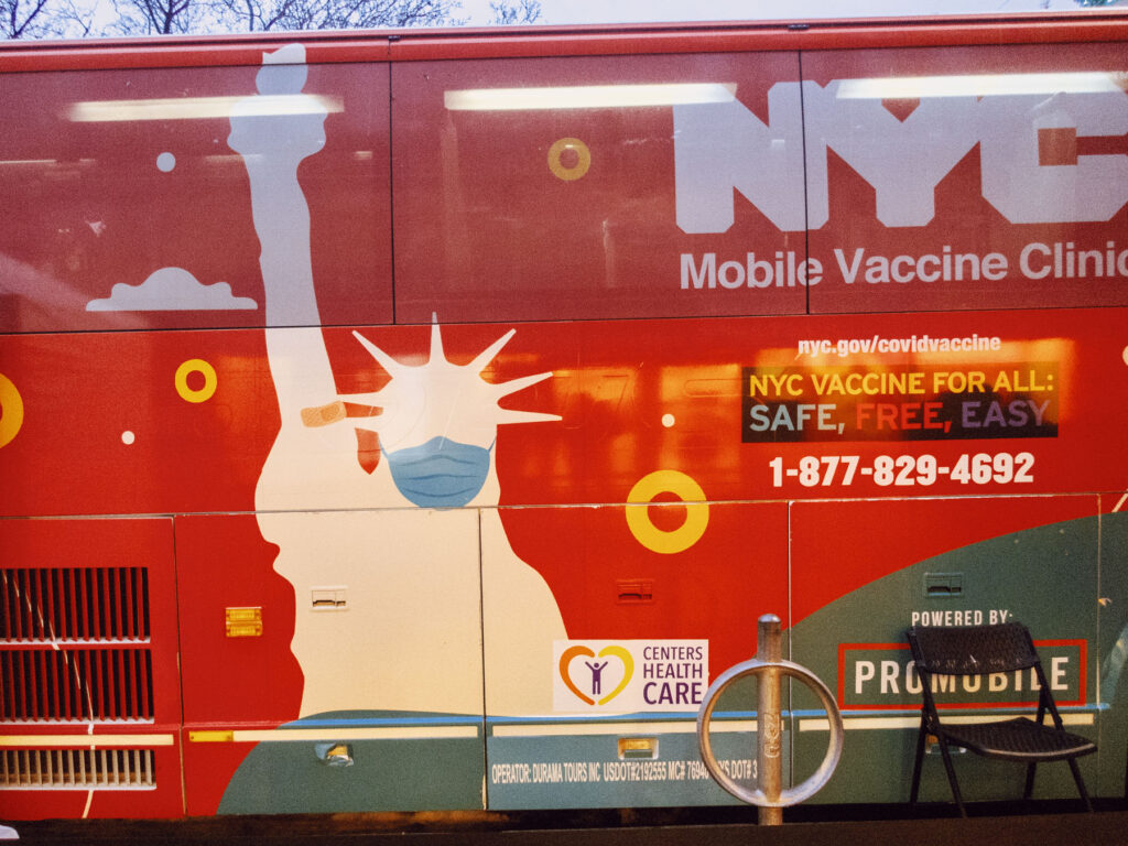 A street photography capture of an NYC Mobile Vaccine Clinic bus, painted in bright red with colorful graphics. The artwork features a stylized Statue of Liberty wearing a face mask and a small bandage on her arm, symbolizing vaccination. Bold text reads 'NYC Vaccine for All: Safe, Free, Easy,' along with a hotline number and website for more information. A folding chair and a bike rack in the foreground add a practical urban touch to the scene.