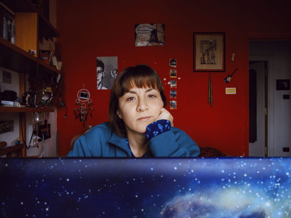 A woman with brown hair, wearing a blue sweater, is sitting in a room with a red wall. She is looking directly at the camera with a neutral expression, resting her chin on her hand. The foreground shows part of a laptop with a galaxy-themed cover. The background includes various decorations and shelves with items.