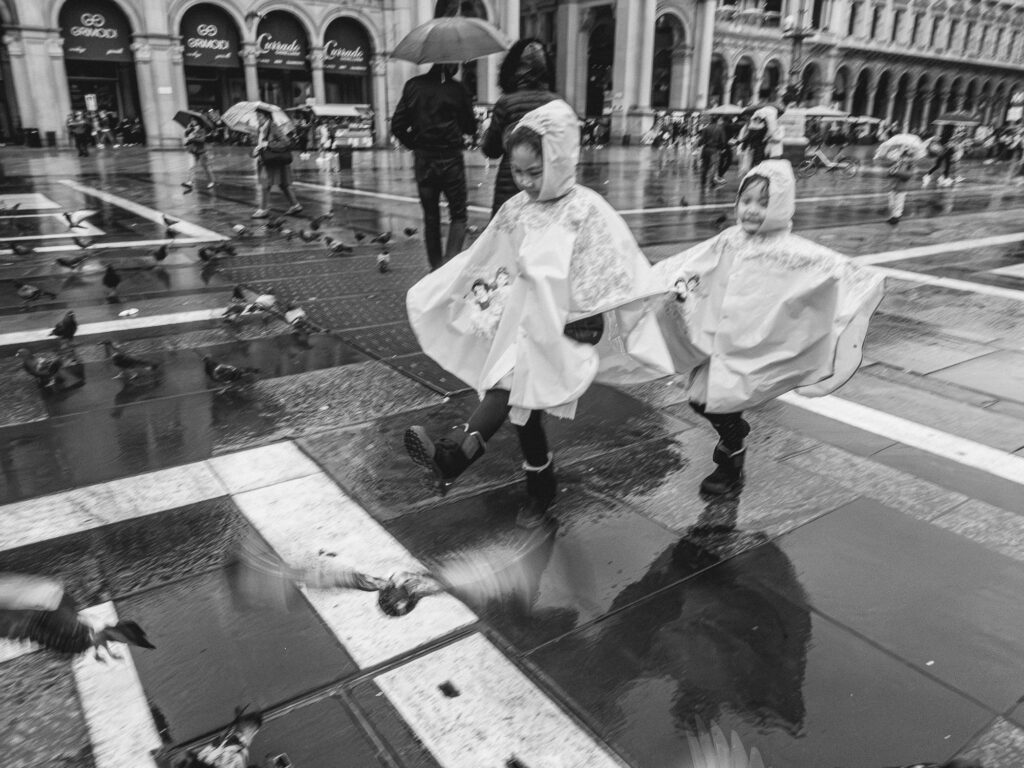 Black and white street photography of a rainy scene in a city square. Two children joyfully run among pigeons, wearing transparent rain ponchos. Pedestrians with umbrellas and scattered birds are in the background, with reflections of the surroundings captured in wet, shiny pavement.