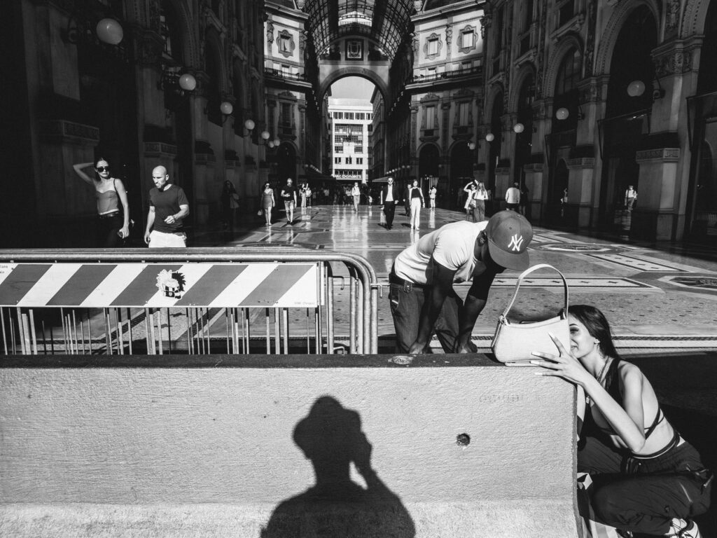 street photography scene in a grand architectural setting, possibly a shopping gallery. A couple walks past in the foreground, while in the bottom right, a woman kneels to speak to a man leaning over a striped barricade. The shadow of the photographer is prominently cast on a wall in the immediate foreground. Background showcases the intricate details of the gallery with arches, decorative floors, and other pedestrians in motion.