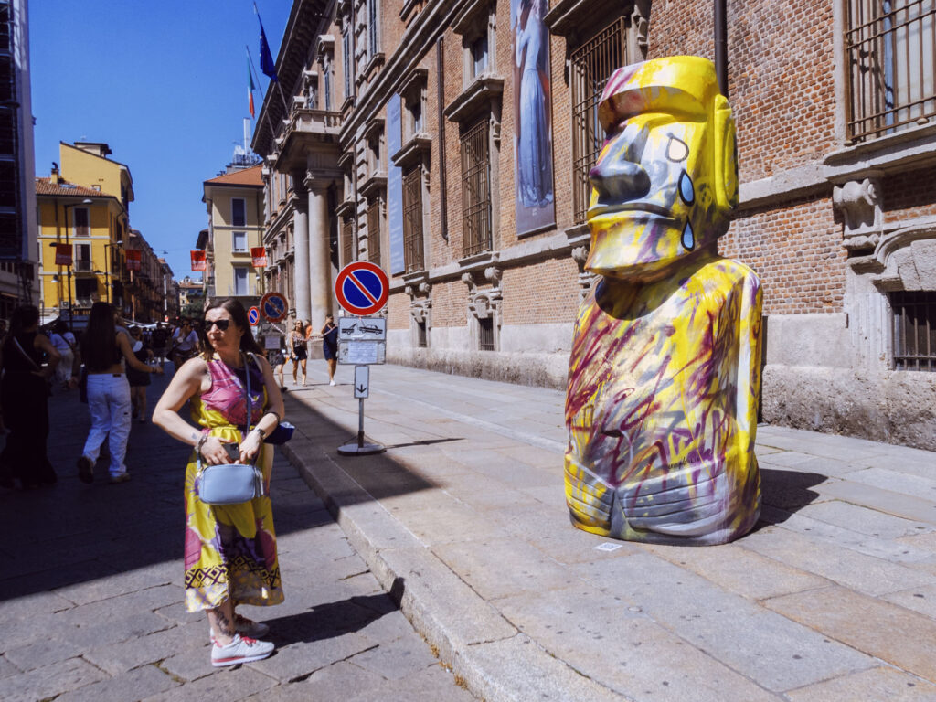 A colorful street scene featuring a woman standing near a large, graffiti-covered statue resembling a Moai head, painted in bright yellow with expressive details, including a teardrop motif. The woman, wearing a vibrant pink and yellow dress, white sneakers, and sunglasses, holds a light blue handbag while looking to her side. Behind her, a historic brick building with large windows and banners lines the street, and a narrow road extends into the distance with more people walking in the sunlit urban environment. Italian flags and no-parking signs add to the bustling atmosphere.