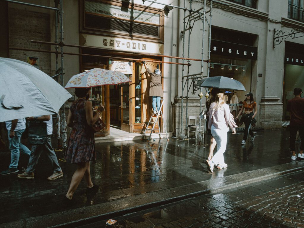 This image captures a bustling street scene during a rain shower. People are navigating a wet cobblestone street with umbrellas in hand, reflecting a diverse range of reactions to the weather. One prominent figure is walking confidently without an umbrella, while others are more cautiously shielded. The scene includes storefronts like "Gay-Odin" and "Sisley," adding an urban commercial feel to the atmosphere. The sunlight peeking through, illuminating raindrops and wet surfaces, creates a lively and dynamic environment, typical of a rainy day in a busy city.