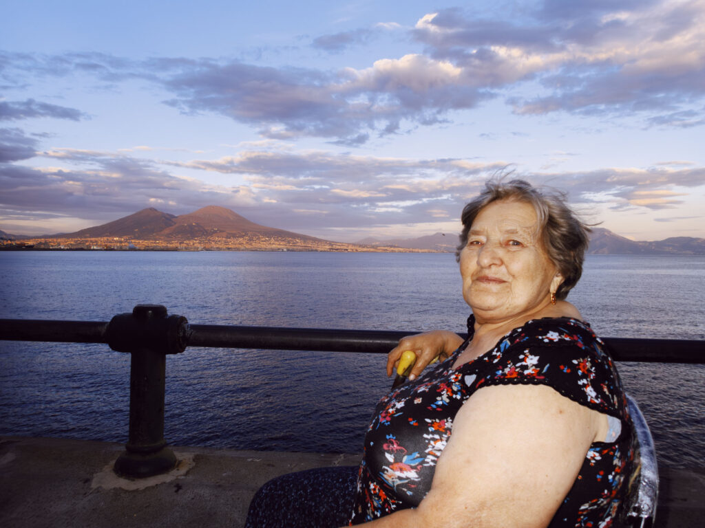 A portrait of an elderly woman sitting along the Naples waterfront during sunset. She is dressed in a floral-patterned top and gazes warmly at the camera, her hand resting on a cane. Behind her, the Gulf of Naples stretches into the distance, with the iconic Mount Vesuvius bathed in golden light under a dramatic sky filled with scattered clouds. The serene water and the warm hues of the scene evoke a peaceful yet lively atmosphere, blending human connection with the grandeur of the natural landscape.