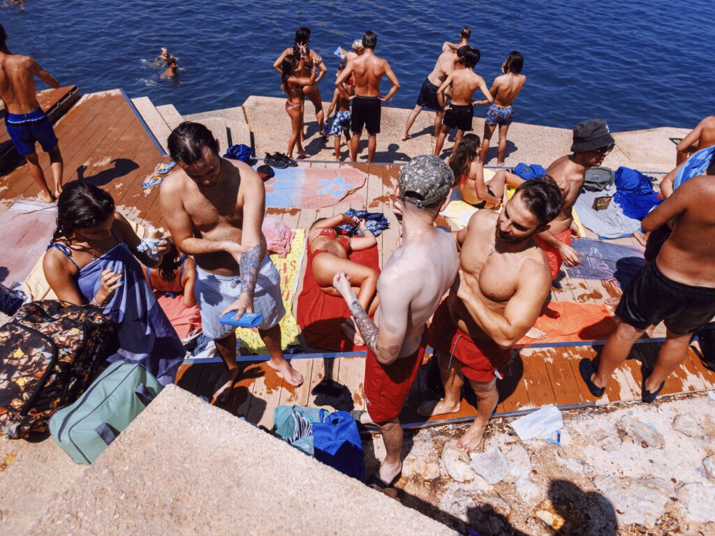 A bustling seaside scene where a group of people enjoys the sun on a wooden pier. Several individuals are sunbathing, while others stand and chat, with a couple of people in the water. The attire is summery—swimwear in various colors and patterns. Beach towels and bags are scattered around, adding to the casual, leisurely atmosphere on a bright sunny day.